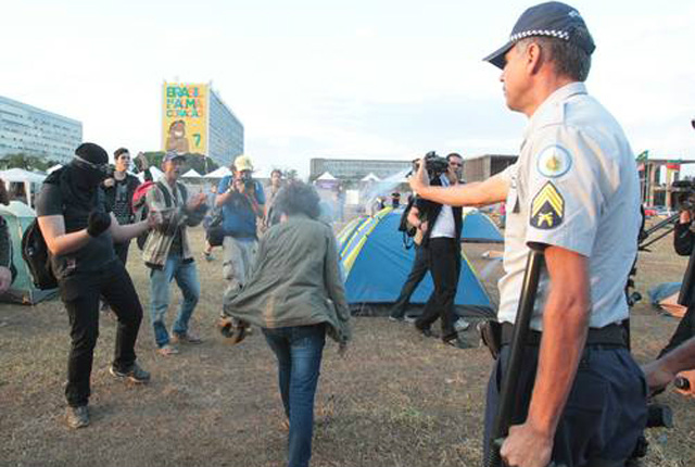 Um policial militar aplica spray de pimenta em uma manifestante, enquanto outro do protesto, encapuzado e com punhos armados ameaça reação (Foto: Google)