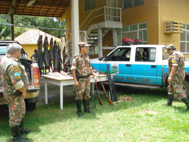 Policiais ambientais exibem armas apreendidas e no fundo os animais abatidos ilegalmente (Foto: PMA)